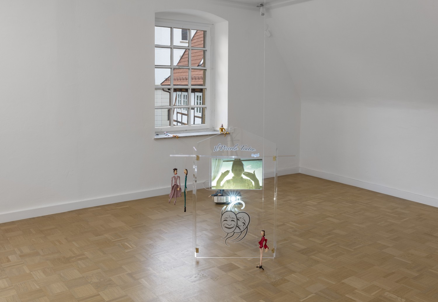 Die Arbeit "Il piccolo teatro" von Julie Béna stellt ein Puppentheater aus Plexiglas dar, das auf dem Boden steht. Auf das Objekt wird ein Video projiziert, das eine Handperformance der Künstlerin zeigt. Im Raum, um das Objekt herum, sind kleine Marionettenpuppen platziert.